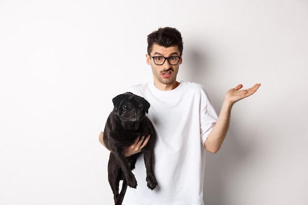 Image d'un beau jeune homme tenant un carlin noir et ayant l'air confus. Guy haussant les épaules et regardant indécis à la caméra, porter le chien dans les bras, fond blanc