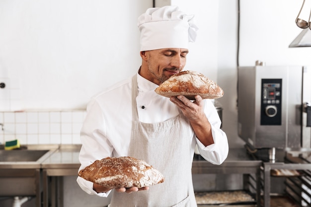 Image de beau mâle boulanger en uniforme blanc debout à la boulangerie, et tenant du pain