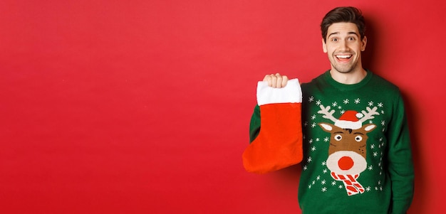 Image de beau mec heureux en pull tenant des bas de Noël et souriant amusé debout contre...