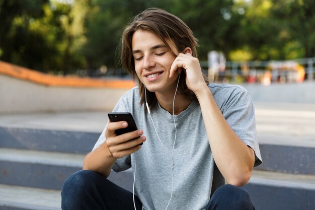 Image de beau mec jeune patineur joyeux assis dans le parc à écouter de la musique avec des écouteurs à l'aide du téléphone.