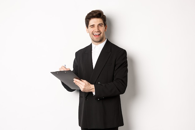 Image d'un beau patron prospère en costume noir, clignotant et souriant tout en signant des documents