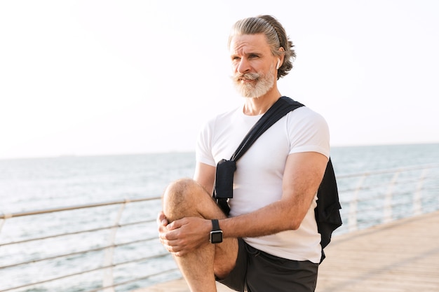 Image d'un beau vieil homme en tenue de sport utilisant un écouteur tout en étirant sa jambe sur la promenade le matin