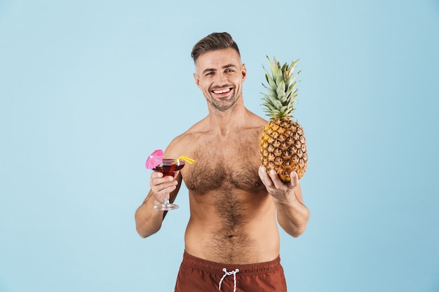 Image d'un bel homme adulte heureux excité posant sur un mur bleu boire un cocktail tenant ananas.