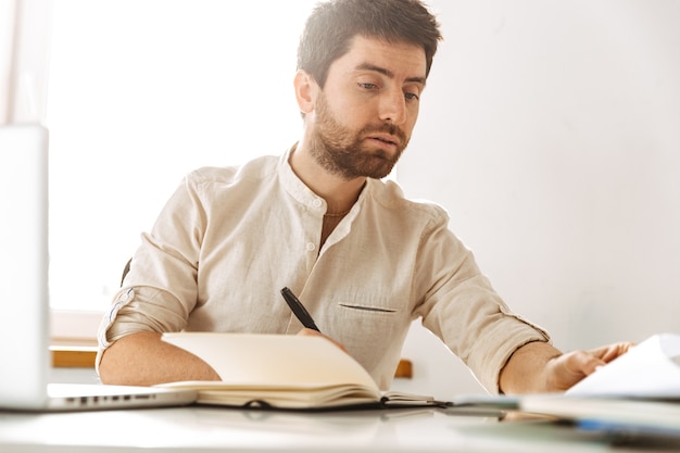 Image de bel homme d'affaires de 30 ans portant une chemise blanche travaillant avec un ordinateur portable et des documents papier, alors qu'il était assis dans un bureau lumineux