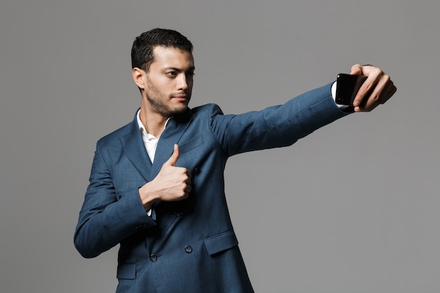 Image d'un bel homme d'affaires prendre un selfie par téléphone mobile posant isolé sur mur de mur gris.