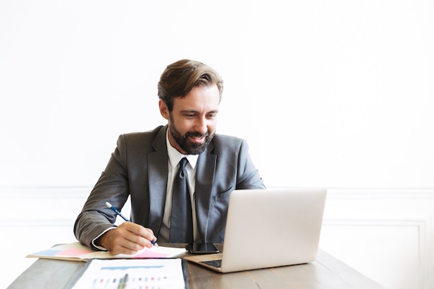 Image d'un bel homme d'affaires souriant portant un costume formel travaillant sur un ordinateur portable au bureau tout en écrivant des notes