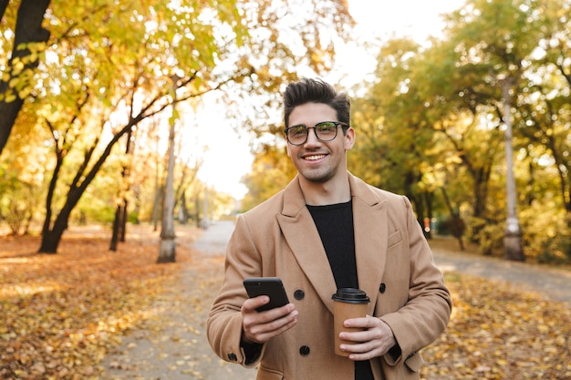 Image d'un bel homme européen utilisant un téléphone portable et souriant en marchant dans un parc en automne