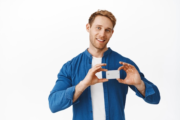 Image d'un bel homme rousse montre une carte de crédit près de son amour de cœur et comme une banque souriante heureuse de recommander une entreprise debout sur fond blanc