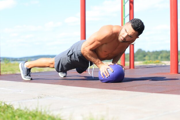 Photo image d'un bel homme sportif fort faisant des exercices avec un ballon de fitness à l'extérieur par une journée ensoleillée.