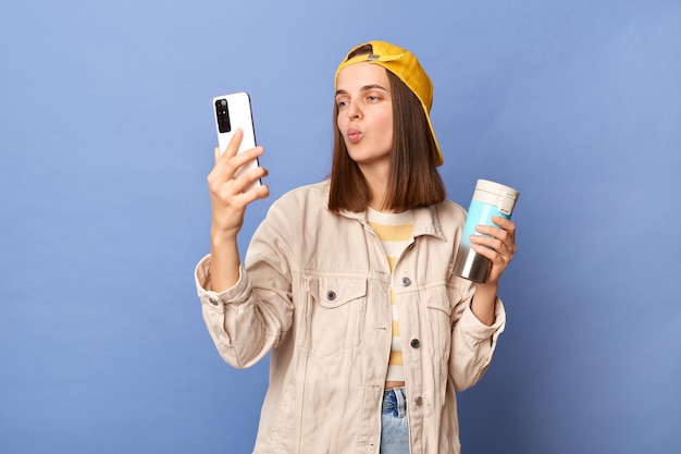 Image d'une belle adolescente caucasienne romantique portant une casquette de baseball et une veste posant isolée sur fond bleu buvant du café ou du thé tenant un téléphone intelligent envoyant un baiser aérien