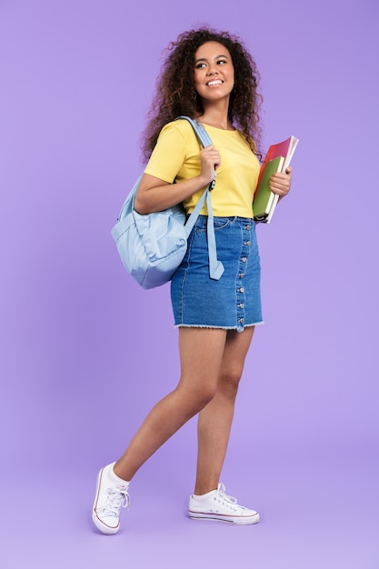 Image d'une belle étudiante afro-américaine aux cheveux bouclés tenant des cahiers d'exercices isolés sur un mur violet