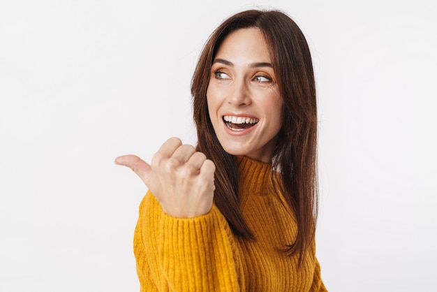 Image de belle femme adulte brune portant un pull souriant et pointant le doigt de côté à copyspace isolated on white
