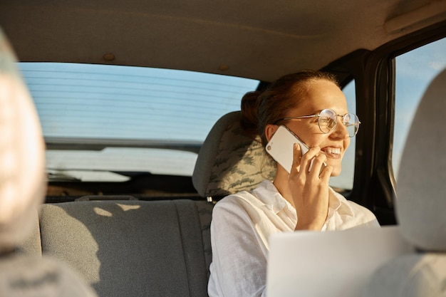 Image D'une Belle Femme D'affaires Joyeuse Assise Sur Le Siège Arrière De La Voiture Et Travaillant Avec Un Ordinateur Portable Et Parlant Sur Un Téléphone Intelligent En Regardant La Fenêtre Du Véhicule