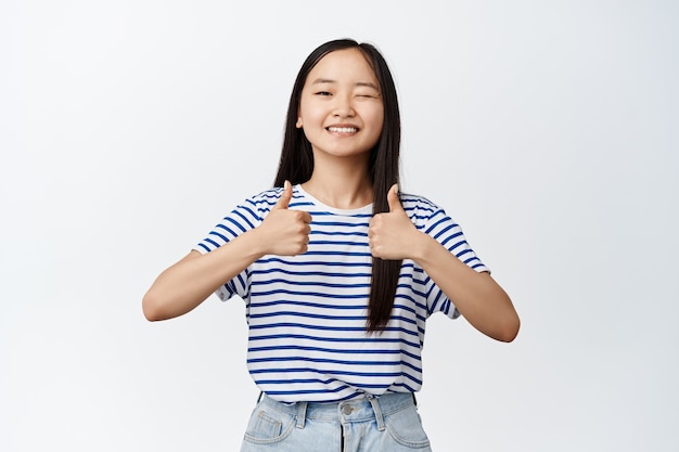 Image d'une belle femme asiatique faisant un clin d'œil et souriant, montrant les pouces vers le haut, approuve et aime le geste, signe de rétroaction positive, debout sur blanc