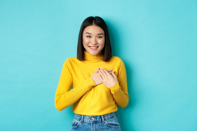 Image de la belle femme asiatique tenant les mains sur le cœur et souriant, vous remerciant, se sentant reconnaissant, debout sur le bleu.