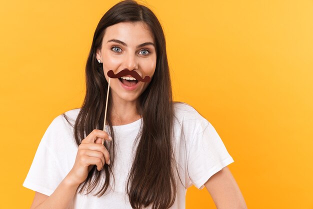 Image d'une belle femme brune portant des vêtements de base s'amusant et tenant une moustache en papier isolée sur un mur jaune