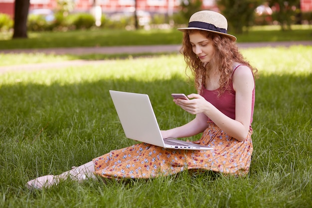 Image de belle femme élégante au chapeau, assis sur l'herbe verte avec un ordinateur portable et un téléphone dans les mains, a des cours en ligne, travaillant en plein air, utilisant Internet sans fil. Concept de pigiste et de style de vie.