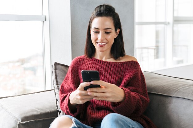 Image d'une belle femme à l'intérieur à la maison sur un canapé à l'aide d'un téléphone mobile.