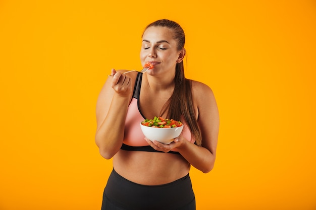 Image de belle femme potelée en survêtement de manger une salade verte de la plaque, isolé sur fond jaune