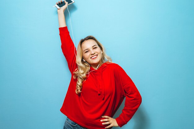 Image d'une belle fille blonde excitée, écouter de la musique dans les écouteurs et danser sur le mur bleu