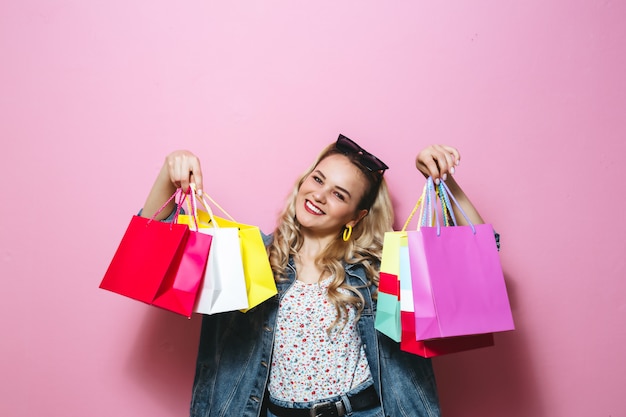 Image d'une belle fille blonde souriante avec des lunettes de soleil, tenant des sacs à provisions sur mur rose