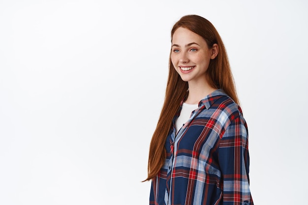 Image d'une belle fille naturelle aux cheveux longs rouges, aspect naturel avec des taches de rousseur et une peau pâle, regardant de côté le logo, des dents blanches souriantes, debout sur fond de studio