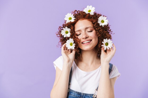 Image d'une belle fille rousse heureuse bouclée posant isolée sur un mur violet avec des fleurs dans les cheveux.