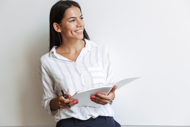 Image d'une belle jeune femme d'affaires positive au bureau isolée sur un mur blanc à l'intérieur en écrivant des notes.