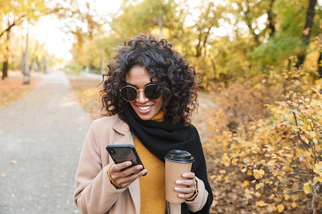 Image d'une belle jeune femme africaine heureuse marchant à l'extérieur dans un parc printanier buvant du café à l'aide d'un téléphone portable.