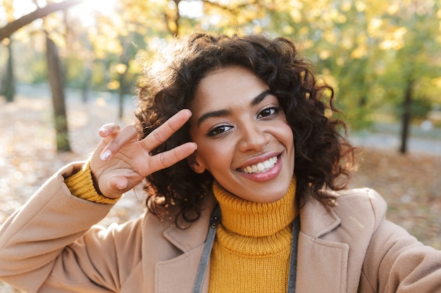 Image d'une belle jeune femme africaine heureuse marchant à l'extérieur dans un parc printanier prendre un selfie par caméra faire un geste de paix.