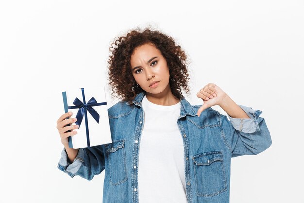 Image d'une belle jeune femme africaine mécontente posant isolée sur un mur blanc tenant une boîte-cadeau présente montrant les pouces vers le bas.