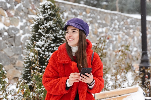 Image de la belle jeune femme à l'extérieur dans la forêt de parc d'hiver de neige à l'aide de téléphone mobile.
