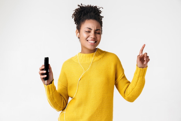 Image d'une belle jeune femme heureuse émotionnelle excitée africaine posant sur un mur blanc, écouter de la musique avec des écouteurs à l'aide de téléphone mobile.