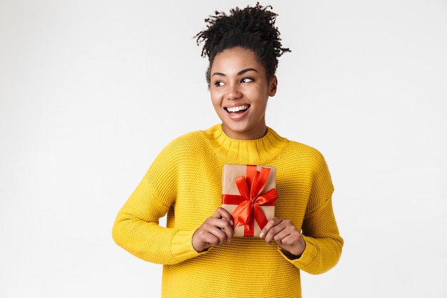 Image d'une belle jeune femme heureuse émotionnelle excitée africaine posant sur un mur blanc tenant une boîte cadeau.