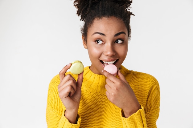 Image d'une belle jeune femme heureuse émotionnelle excitée africaine posant sur un mur blanc tenant des macarons sucrés.