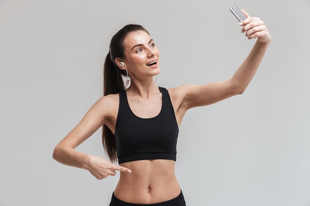 Image d'une belle jeune femme de remise en forme sportive utilisant un téléphone portable écoutant de la musique isolée sur un mur gris prendre un selfie.