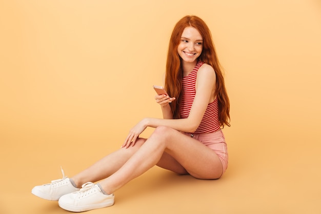 Image d'une belle jeune femme rousse souriante et heureuse posant isolée sur un mur jaune à l'aide d'un téléphone portable.