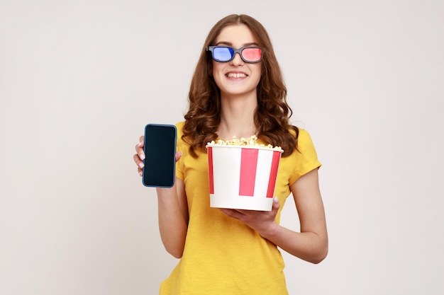 Image d'une belle jeune femme tenant du pop-corn dans un seau, montrant un smartphone avec un écran vide vide, souriant à la caméra, porte un T-shirt jaune. Tourné en studio intérieur isolé sur fond gris.