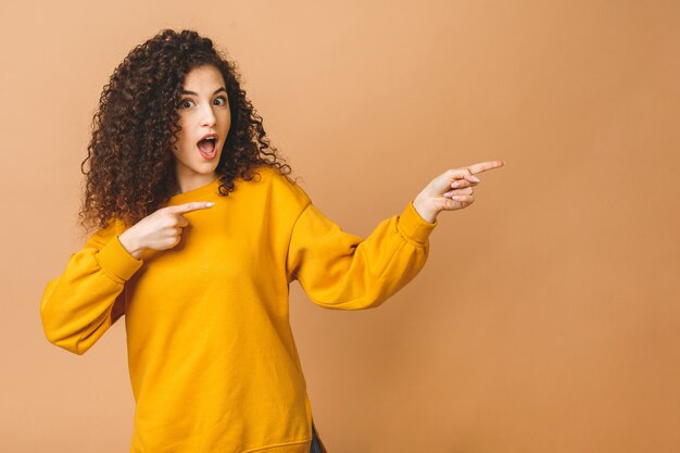 Image de belle jolie jeune femme bouclée debout isolé sur fond beige pointant vers le haut. En regardant de côté.