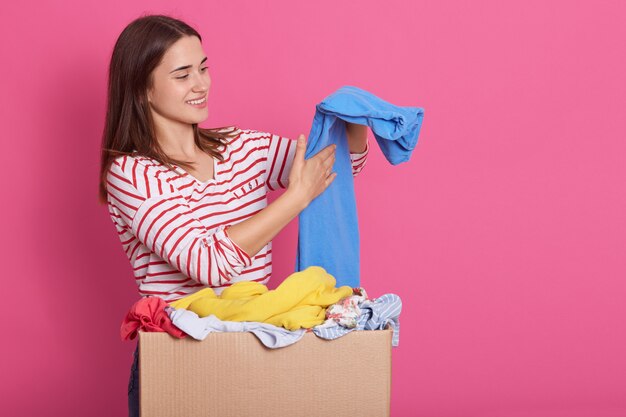 Image de brune séduisante dame regardant de côté, souriant sincèrement, mettant les vêtements donnés dans l'ordre, tenant un pantalon bleu dans les mains