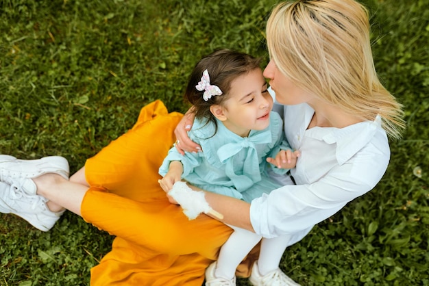 Image candide en plein air d'une belle mère jouant et câlinant avec sa fille profitant du temps ensemble Jolie petite fille embrasse sa mère assise sur l'herbe verte dans le parc Fête des mères