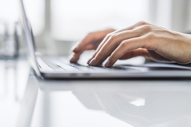 L'image capture des mains féminines dans les moindres détails en train de taper sur un clavier d'ordinateur portable élégant avec un environnement de bureau flou en arrière-plan