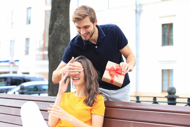 Image d'un charmant couple excité en vêtements d'été souriant et tenant une boîte présente ensemble tout en étant assis sur un banc.