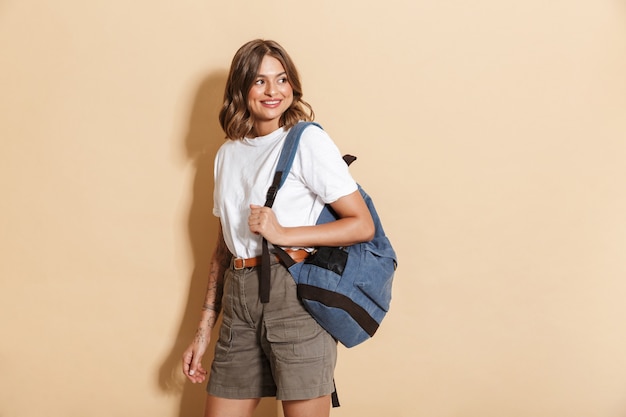 Image d'une charmante adolescente portant un sac à dos souriant et marchant isolée sur un mur beige