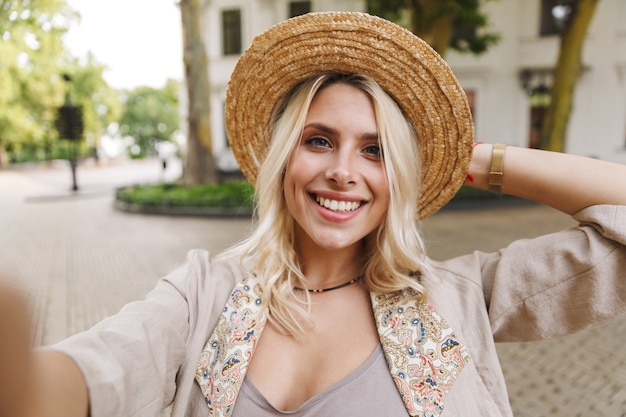 Image de charmante femme portant costume et chapeau de paille souriant, tout en prenant selfie photo en plein air au centre-ville