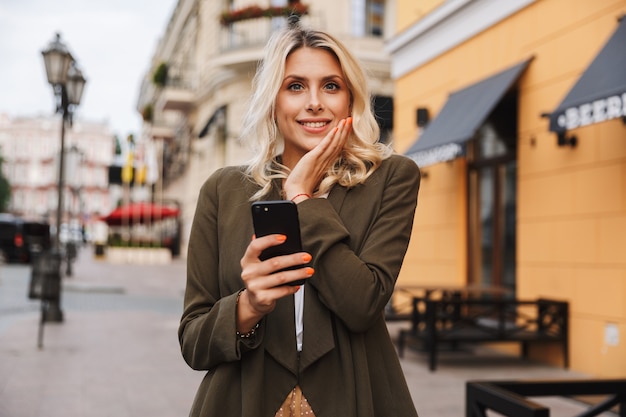 Image de charmante femme souriante et à l'aide de téléphone portable, tout en marchant dans la rue de la ville