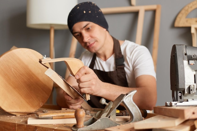 Image d'un charpentier calme et séduisant jeune homme adulte portant un t-shirt blanc, une casquette noire et un tablier marron travaillant dans la menuiserie, fabriquant une chaise en bois, ponçant un bloc de bois