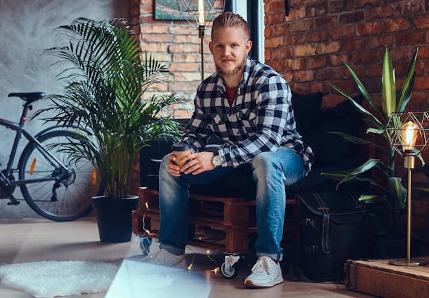 L'image complète du corps d'un homme hipster blond boit du café dans une pièce avec un intérieur loft.