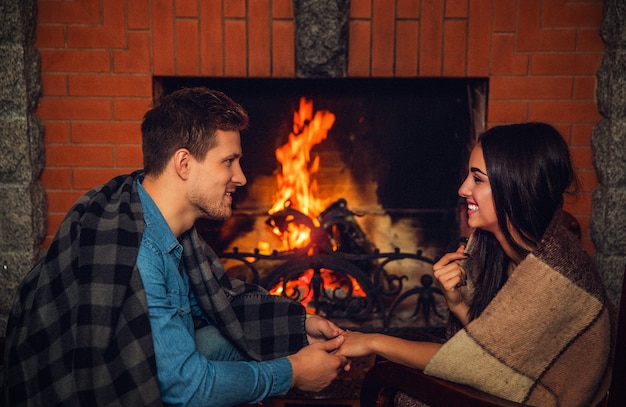 Image confortable de charmant jeune homme et femme se regardent et sourient. Ils ont des couvertures sur les épaules. Jeune couple, s'asseoir cheminée, à, fire.