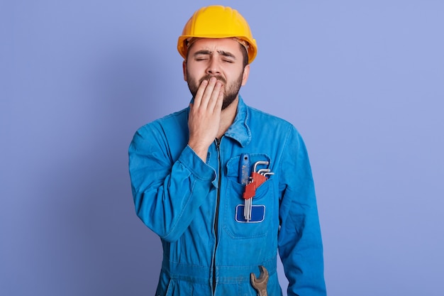 Image d'un constructeur épuisé et somnolent qui bâille, se couvrant la bouche avec la main, fermant les yeux, ayant la barbe, portant l'uniforme et le casque, étant fatigué du travail, ayant envie de dormir. Concept de personnes et de travail.